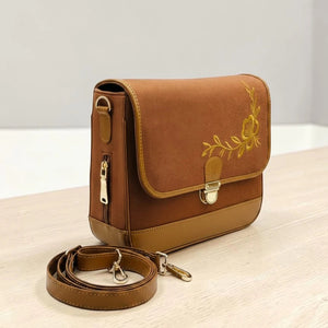 Needlesay botanica cocoa a brown denim crossbody bag with a tan leather base, featuring a yellow floral embroidery on the front flap and a matching adjustable shoulder strap, displayed on a light wooden surface.