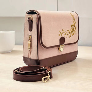 A Needlesay Botanica Blush shoulder bag with gold floral embroidery on the front flap, shown on a light wood tabletop with its matching brown strap placed in front. A white cup is softly blurred in the background.