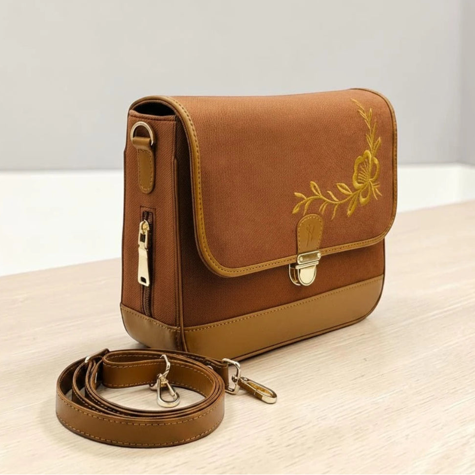Needlesay botanica cocoa a brown denim crossbody bag with a tan leather base, featuring a yellow floral embroidery on the front flap and a matching adjustable shoulder strap, displayed on a light wooden surface.